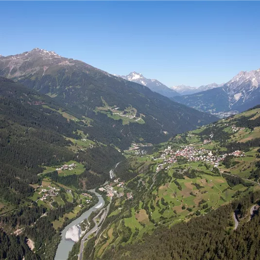 Eine beeindruckende Berglandschaft mit einem klaren blauen Himmel. Ein Fluss schlängelt sich durch das grüne Tal, umgeben von hohen Bergen.