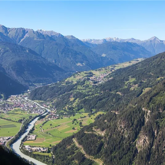 Eine malerische Berglandschaft mit grünen Tälern und einem klaren Fluss. Im Hintergrund sind schneebedeckte Gipfel und ein blauer Himmel zu sehen.