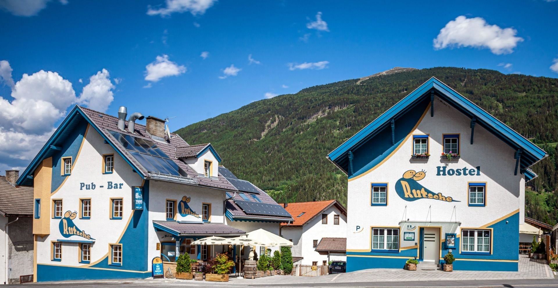 Ein buntes Hostel mit einem Pub und einer Bar in einer malerischen Berglandschaft. Die Gebäude sind freundlich gestaltet und laden zum Verweilen ein.