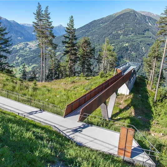 Eine moderne Aussichtsplattform in den Bergen, umgeben von grünen Wiesen und Nadelbäumen. Im Hintergrund sind majestätische Berge zu sehen.