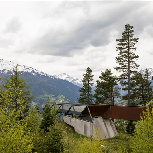 Eine moderne Aussichtsplattform umgeben von Bäumen, mit Blick auf die Berge. Der Himmel ist bewölkt, und die Landschaft zeigt eine Mischung aus Grün und schneebedeckten Gipfeln.