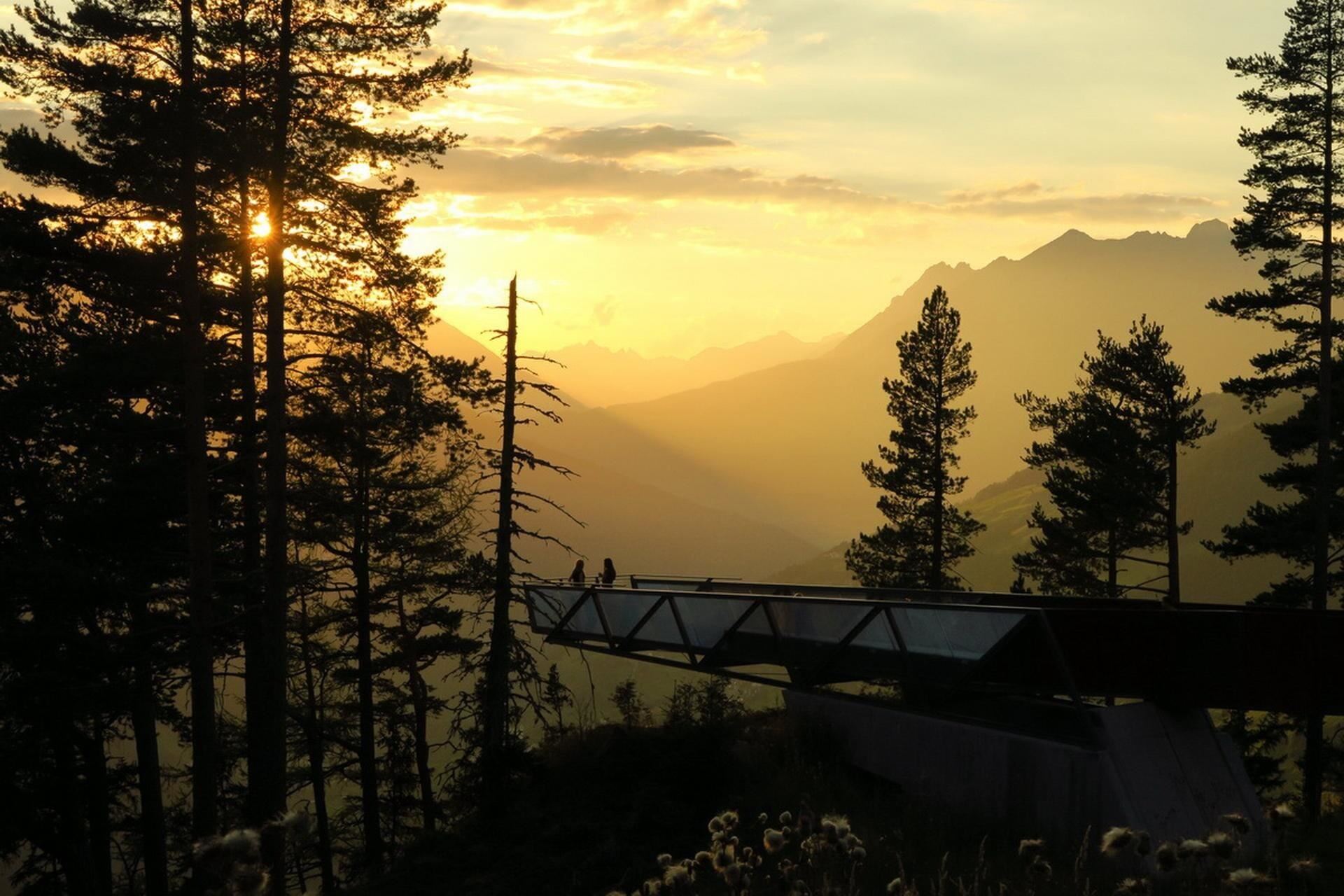 Ein malerischer Sonnenuntergang hinter Bergen, umgeben von Bäumen. Zwei Personen genießen die Aussicht auf einer Aussichtsplattform.