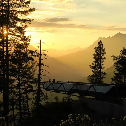 Ein malerischer Sonnenuntergang hinter Bergen, umgeben von Bäumen. Zwei Personen genießen die Aussicht auf einer Aussichtsplattform.