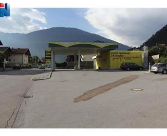 Eine moderne Tankstelle mit einem hellen Dach in einer ländlichen Umgebung. Im Hintergrund sind Berge und Wohnhäuser zu sehen.