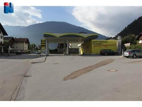 Eine moderne Tankstelle mit einem hellen Dach in einer ländlichen Umgebung. Im Hintergrund sind Berge und Wohnhäuser zu sehen.