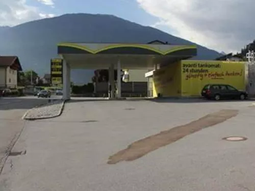 Eine moderne Tankstelle mit einem hellen Dach in einer ländlichen Umgebung. Im Hintergrund sind Berge und Wohnhäuser zu sehen.