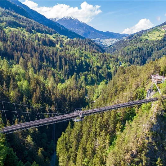 An impressive suspension bridge over a deep valley, surrounded by lush forests and mountains. The bright blue sky provides a picturesque view.