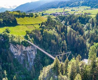 Eine spektakuläre Hängebrücke über eine Schlucht, umgeben von dichten Wäldern und grünen Wiesen. Im Hintergrund sind sanfte Hügel und Berge zu sehen.