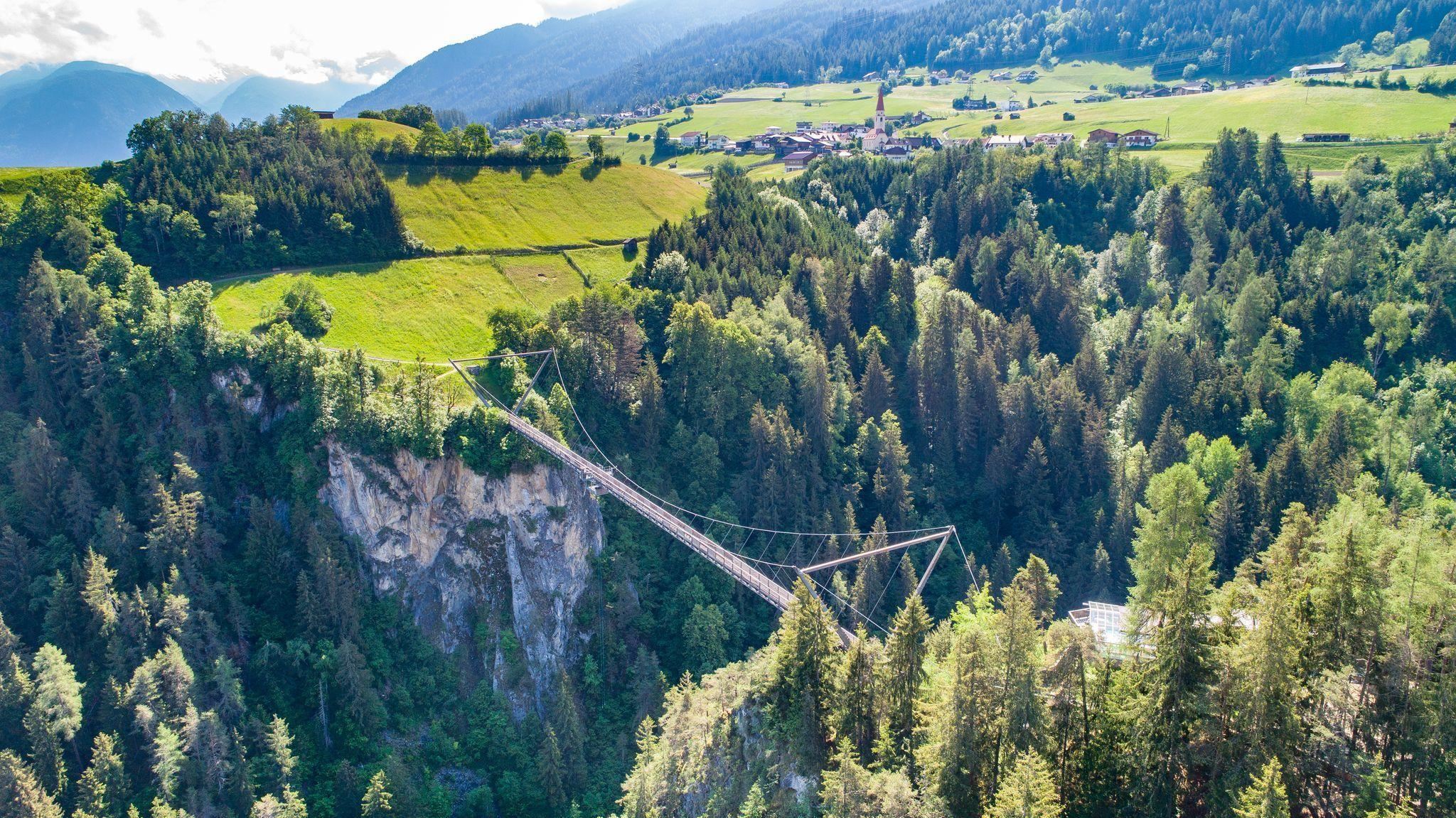 An impressive suspension bridge over a gorge, surrounded by dense forests and gentle hills. In the background, green fields and a small village can be seen.