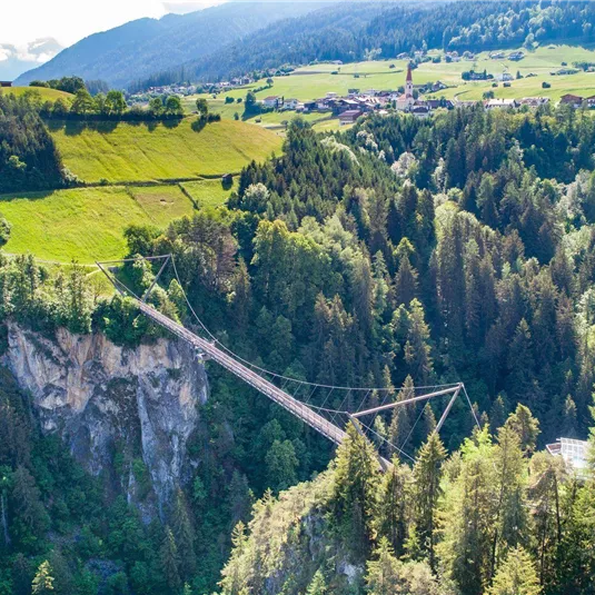 An impressive suspension bridge over a gorge, surrounded by dense forests and gentle hills. In the background, green fields and a small village can be seen.