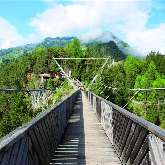 A wooden bridge spans a gorge, surrounded by green trees and mountains. The sky is blue with some clouds.