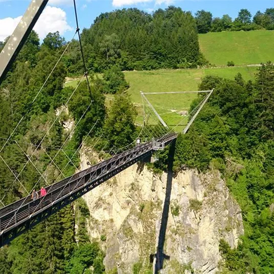 An impressive suspension bridge over a green landscape and a deep chasm. The bridge connects two sides of the valley and offers a picturesque view of the surroundings.