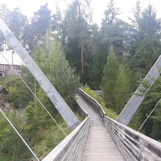 A swinging wooden bridge leads through a green landscape with trees. In the background, a wooden building can be seen.
