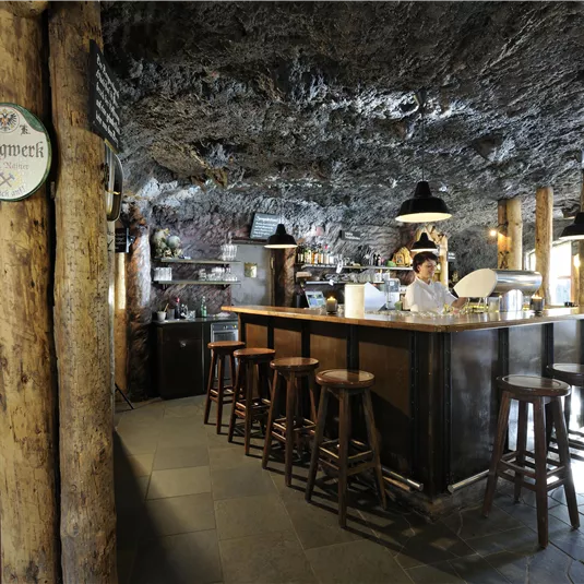 A cozy bar in a cave, with a wooden counter and high stools. Natural rock formations are visible on the wall, creating a unique atmosphere.