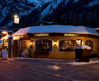 Ein gemütliches Restaurant im Schnee mit warmem Licht und festlicher Dekoration. Im Hintergrund sind schneebedeckte Berge und eine winterliche Landschaft zu sehen.