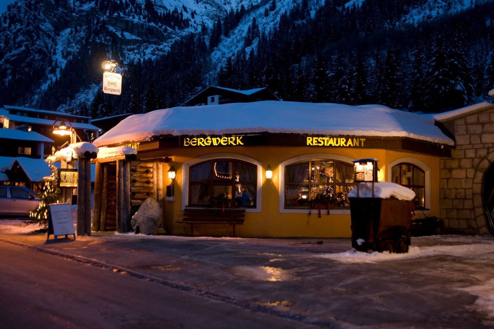 A cozy restaurant in the snow with warm light and festive decorations. In the background, snow-covered mountains and a winter landscape can be seen.