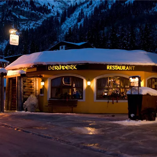 A cozy restaurant in the snow with warm light and festive decorations. In the background, snow-covered mountains and a winter landscape can be seen.