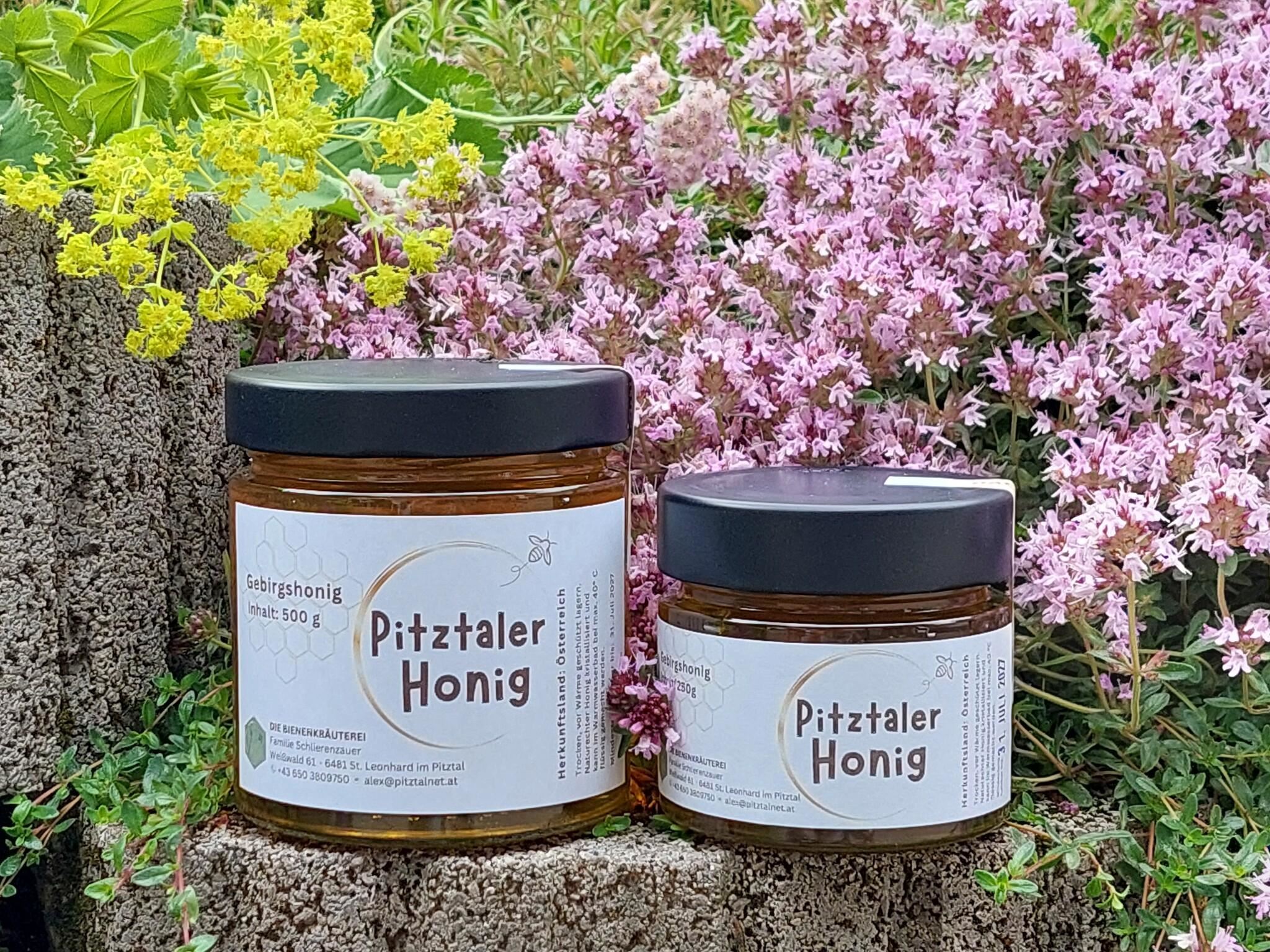 Zwei Gläser Pitztaler Honig stehen vor einer blühenden Blumenlandschaft. Die Gläser sind mit einem schwarzen Deckel versehen und tragen ein einfaches, ansprechendes Etikett.