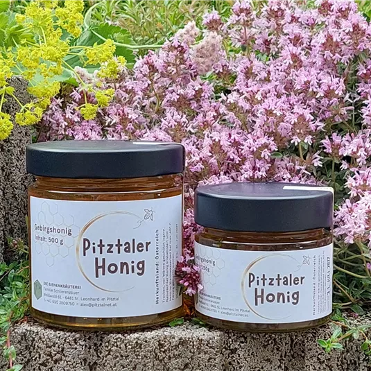 Zwei Gläser Pitztaler Honig stehen vor einer blühenden Blumenlandschaft. Die Gläser sind mit einem schwarzen Deckel versehen und tragen ein einfaches, ansprechendes Etikett.