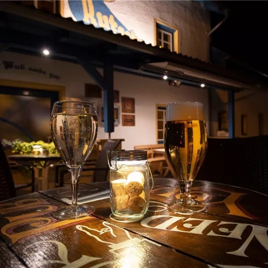 Ein Tisch im Freien mit einem Bier und einem Glas Wein, umgeben von stimmungsvollem Licht. Im Hintergrund ist ein Restaurant mit einer einladenden Atmosphäre zu sehen.