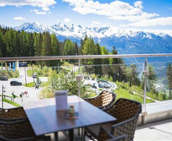 A beautiful view of snow-covered mountains and green forests. On a balcony, there are cozy chairs and a table.