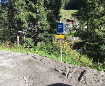 A parking sign stands between trees in a mountainous landscape. In the background, some buildings and a lot of green vegetation can be seen.