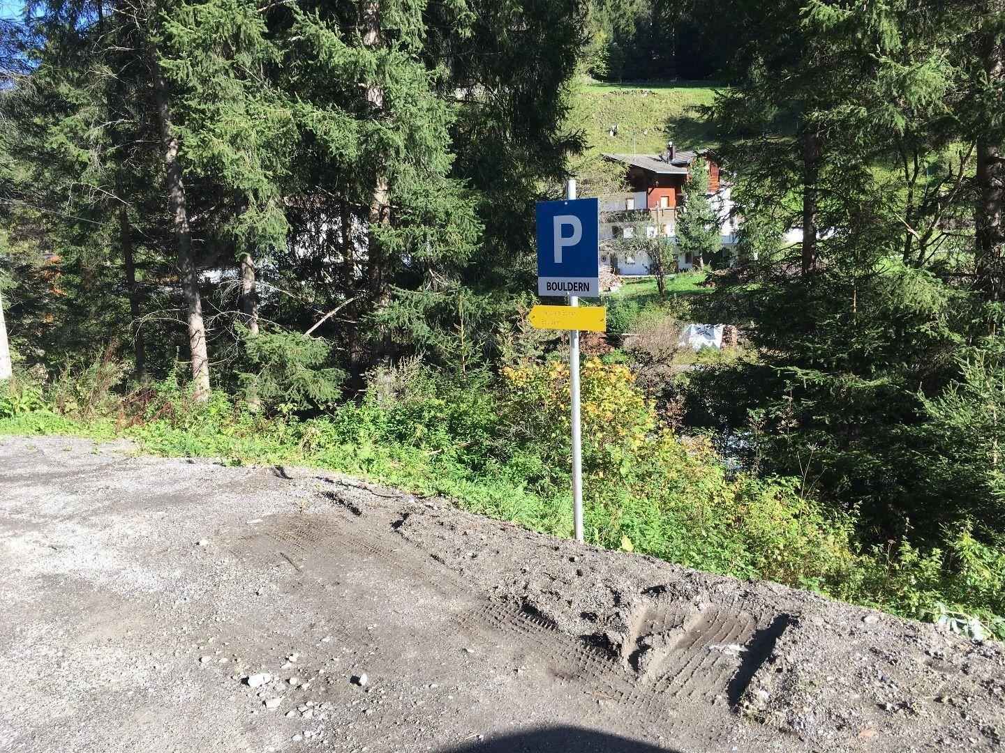 A parking sign stands between trees in a mountainous landscape. In the background, some buildings and a lot of green vegetation can be seen.