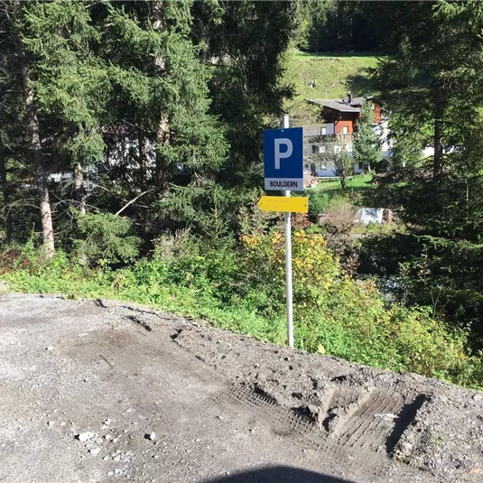 A parking sign stands between trees in a mountainous landscape. In the background, some buildings and a lot of green vegetation can be seen.
