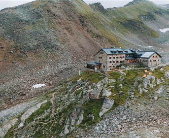 Ein Berggasthaus liegt malerisch auf einem schroffen Hang. Im Hintergrund sind die beeindruckenden, grünen Berge zu sehen.