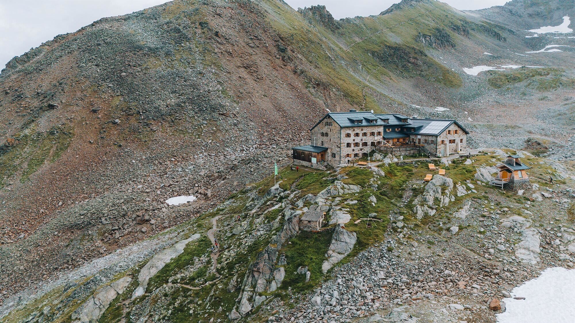 Ein Berggasthaus liegt malerisch auf einem schroffen Hang. Im Hintergrund sind die beeindruckenden, grünen Berge zu sehen.