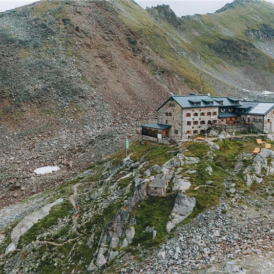 Eine alpine Hütte umgeben von Berglandschaften und felsigem Gelände. Über der Hütte liegt ein bewölkter Himmel.