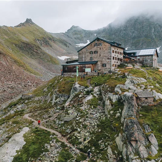 Eine Berghütte liegt auf einem steilen Hang umgeben von Felsen und Bergen. Der Himmel ist bewölkt und eine Wanderweg führt zur Hütte.