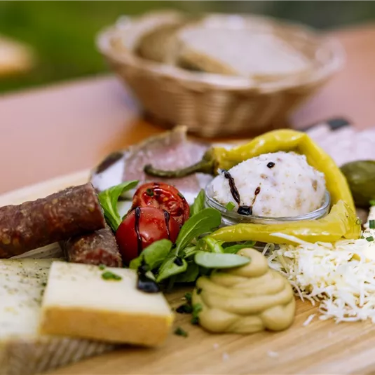 Ein Holzteller mit verschiedenen Käsesorten, Wurst, eingelegtem Gemüse und einer Scheibe Brot. Dazu gehören frische Kräuter und eine kleine Schüssel mit Dip.