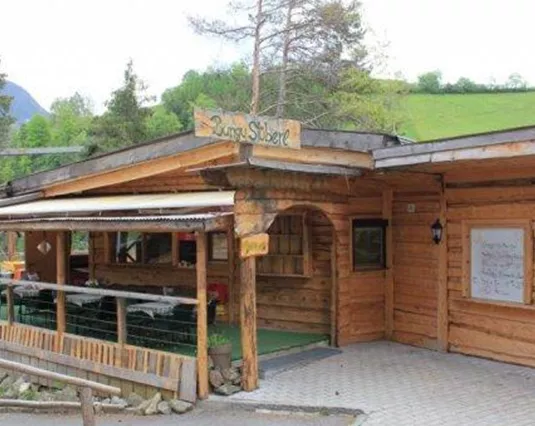 A rustic wooden cabin surrounded by green meadows and mountains. The entrance area is inviting with a covered seating area.