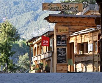 A rustic building with a wooden facade and a sign "Bungy Stube". In the foreground, cozy seating areas and a board with drinks are visible.
