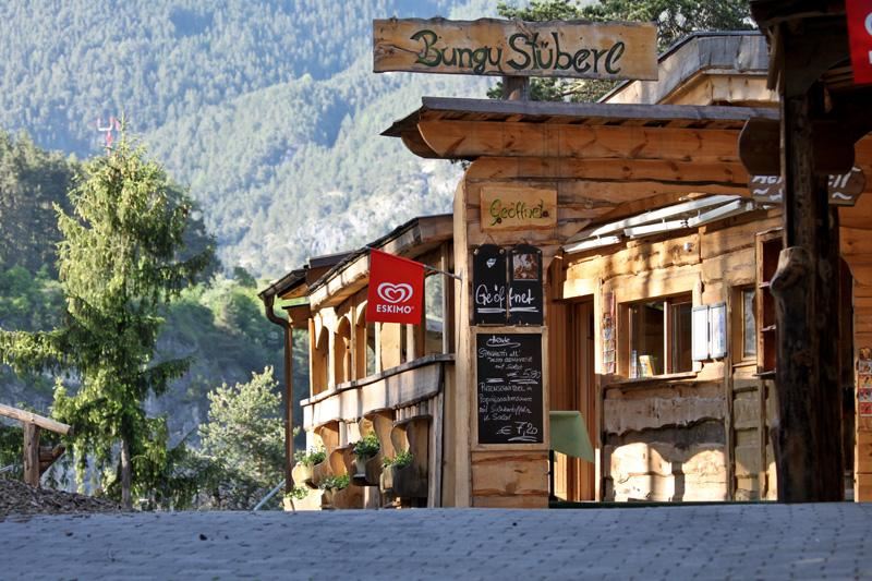A rustic building with a wooden facade and a sign "Bungy Stube". In the foreground, cozy seating areas and a board with drinks are visible.