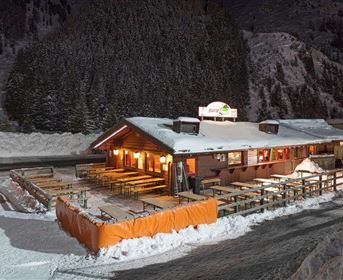 Ein gemütliches Restaurant im Schnee bei Nacht, umgeben von verschneiten Bäumen. Die beleuchtete Fassade und die Terrasse laden zum Verweilen ein.