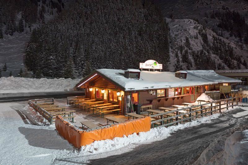 Ein gemütliches Restaurant im Schnee bei Nacht, umgeben von verschneiten Bäumen. Die beleuchtete Fassade und die Terrasse laden zum Verweilen ein.
