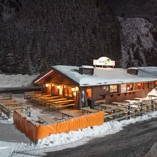 Ein gemütliches Restaurant im Schnee bei Nacht, umgeben von verschneiten Bäumen. Die beleuchtete Fassade und die Terrasse laden zum Verweilen ein.