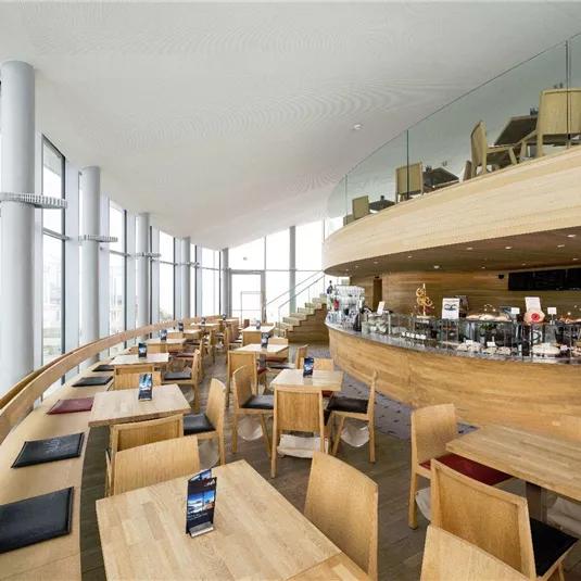 A modern restaurant with wooden furniture and large windows. The atmosphere is bright and welcoming, ideal for social gatherings.