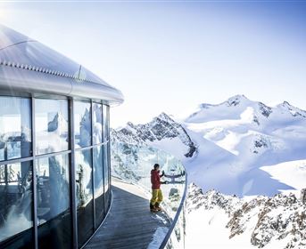 A modern observation platform in the mountains with snow-capped peaks in the background. A person stands by the railing and enjoys the view.