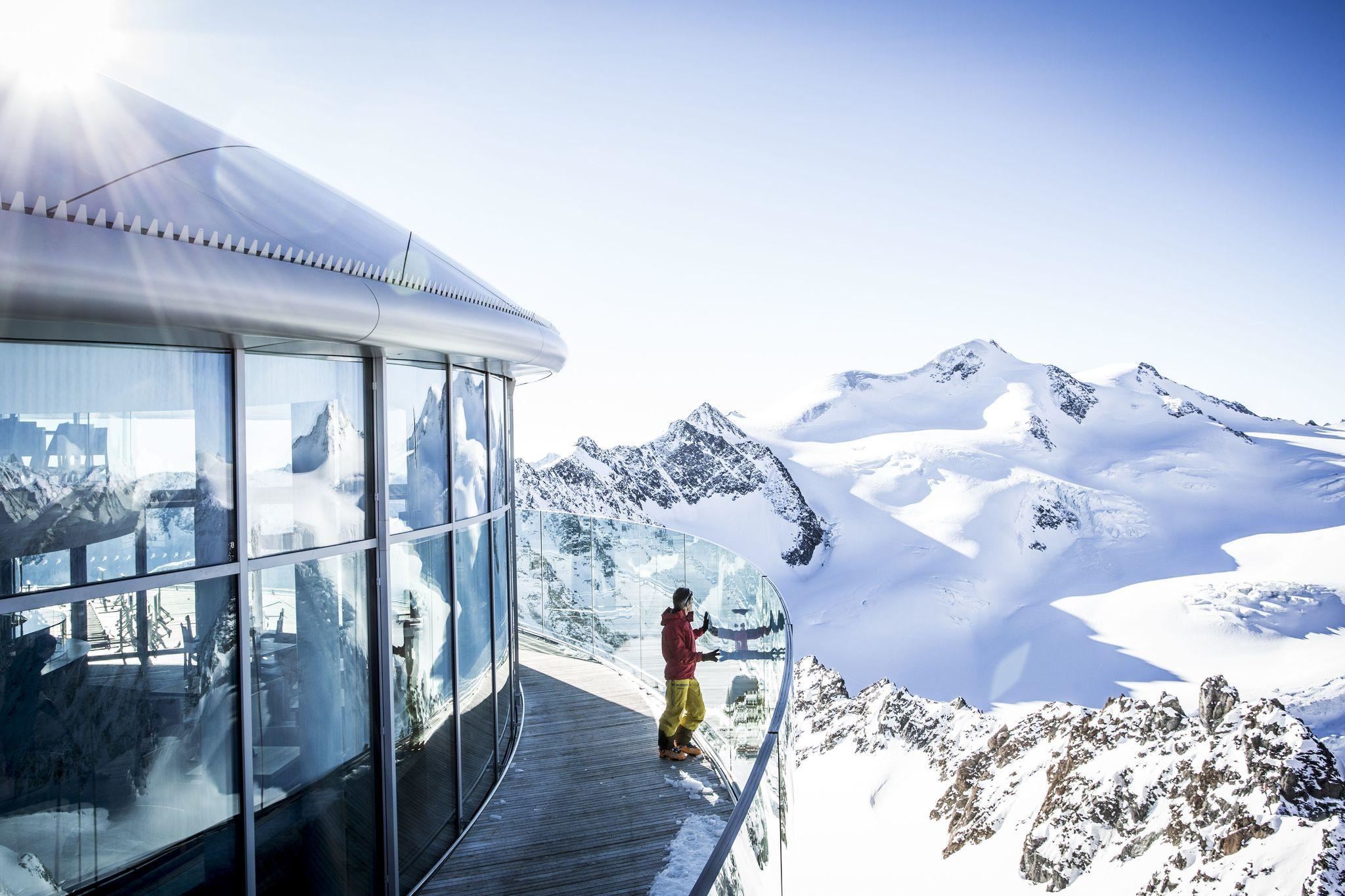 A modern observation platform in the mountains with snow-capped peaks in the background. A person stands by the railing and enjoys the view.