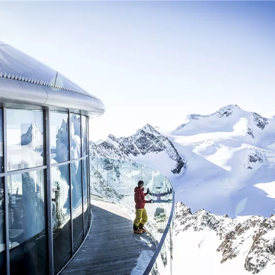 A modern observation platform in the mountains with snow-capped peaks in the background. A person stands by the railing and enjoys the view.