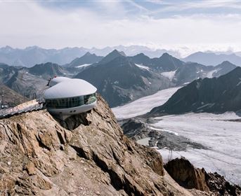 Ein futuristisches Gebäude auf einem Berggipfel mit beeindruckenden Ausblick auf die umliegenden Berge und Gletscher. Die Landschaft ist atemberaubend und dramatisch.