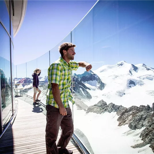 A man stands on a terrace with a glass railing, looking out at an impressive mountain landscape. In the background, there are snow-capped peaks and a blue sky.