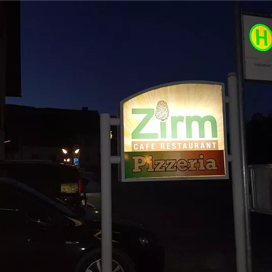 A illuminated sign reading "Zirm Pizzeria" at night. In the background, a bus stop is visible.