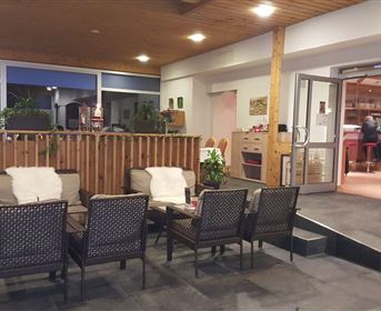 A cozy seating area with armchairs and plants. The room has large windows and a warm wooden ceiling.