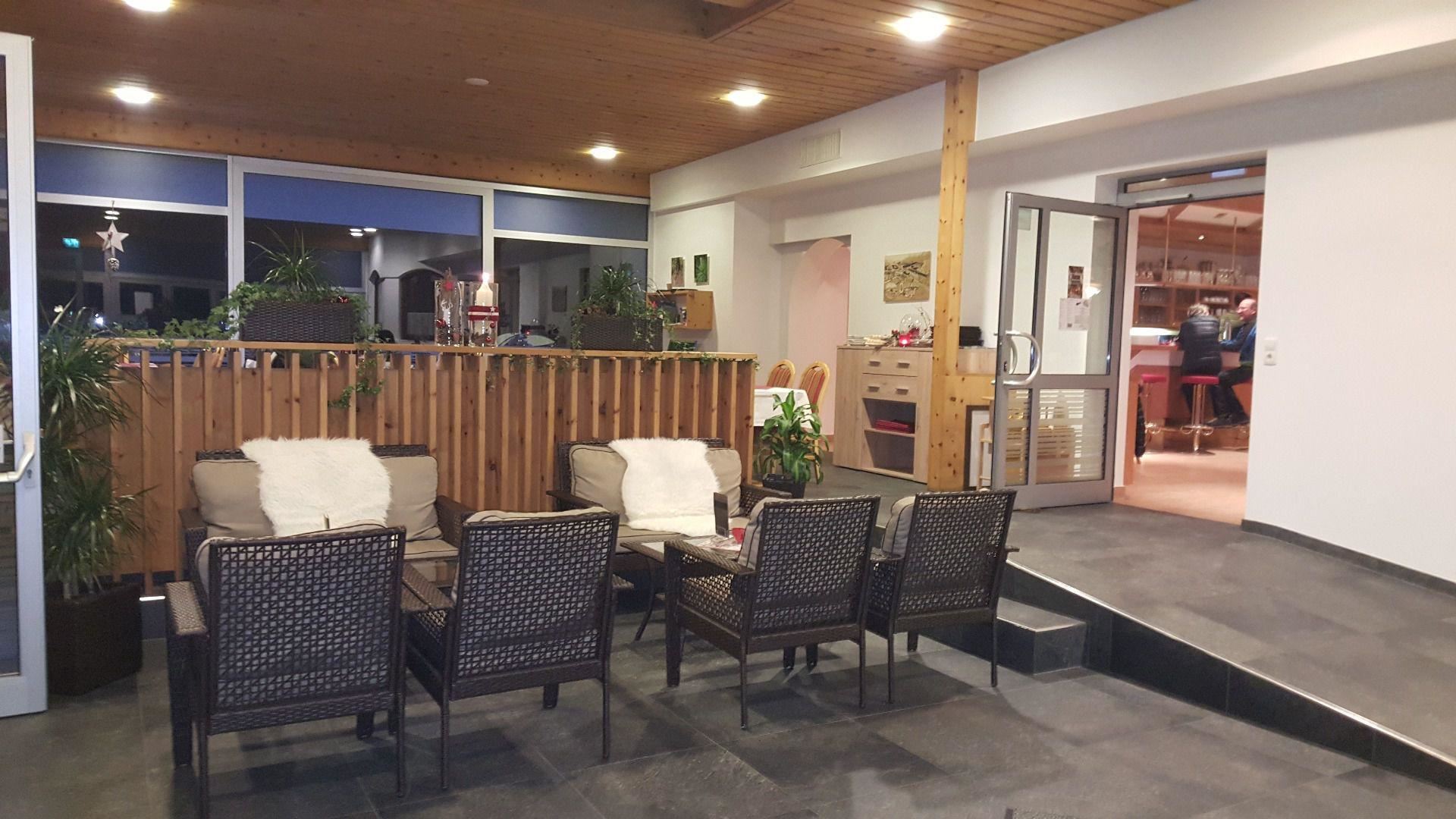 A cozy seating area with armchairs and plants. The room has large windows and a warm wooden ceiling.