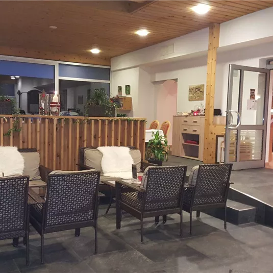 A cozy seating area with armchairs and plants. The room has large windows and a warm wooden ceiling.
