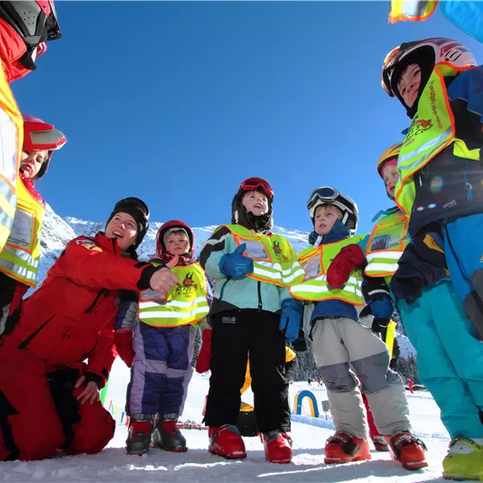 Eine Gruppe von Kindern in bunten Skianzügen versammelt sich im Schnee. Die Skilehrer geben ihnen Anweisungen unter einem klaren blauen Himmel.